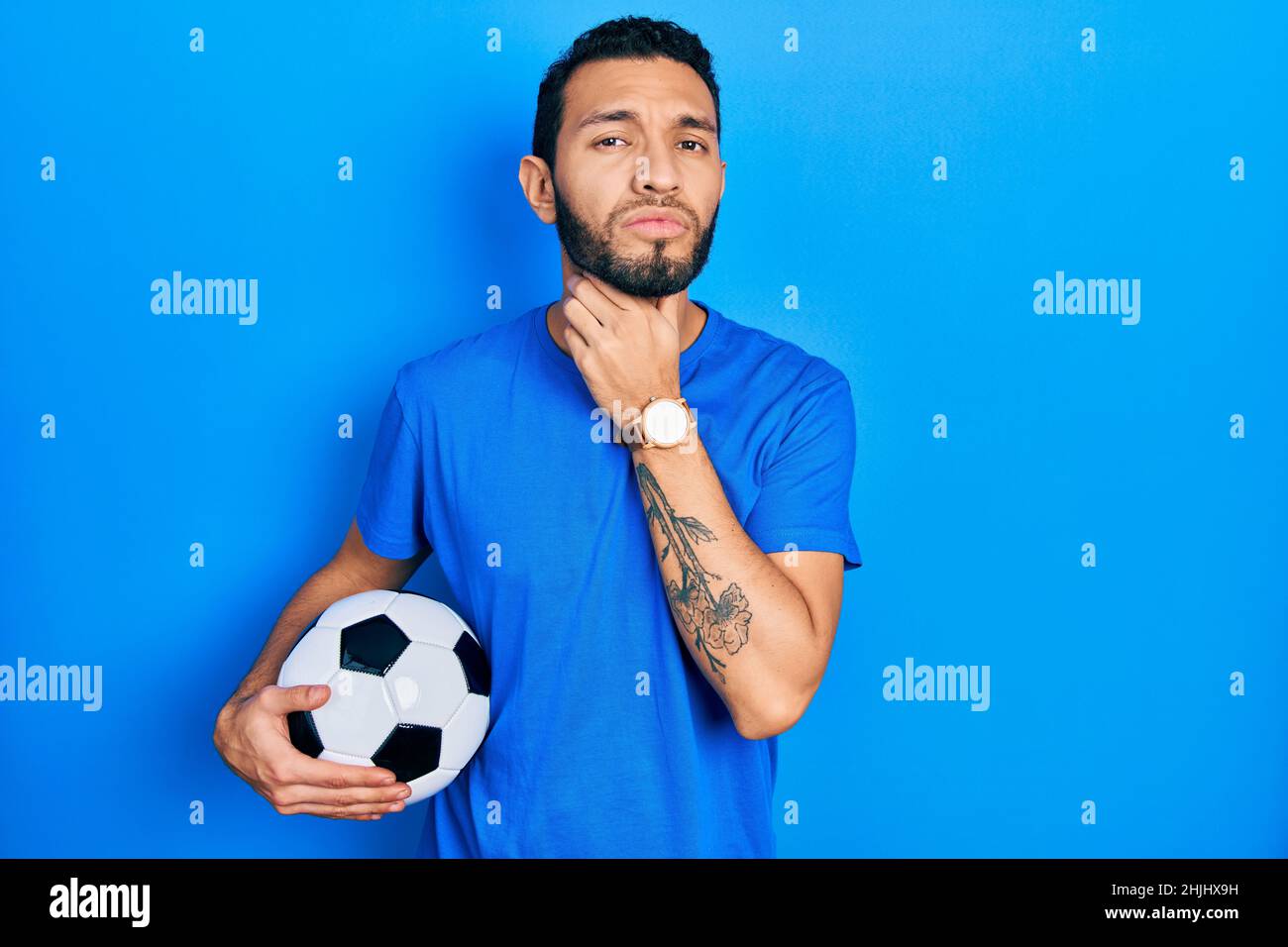 Hispanic man with beard holding soccer ball touching painful neck, sore