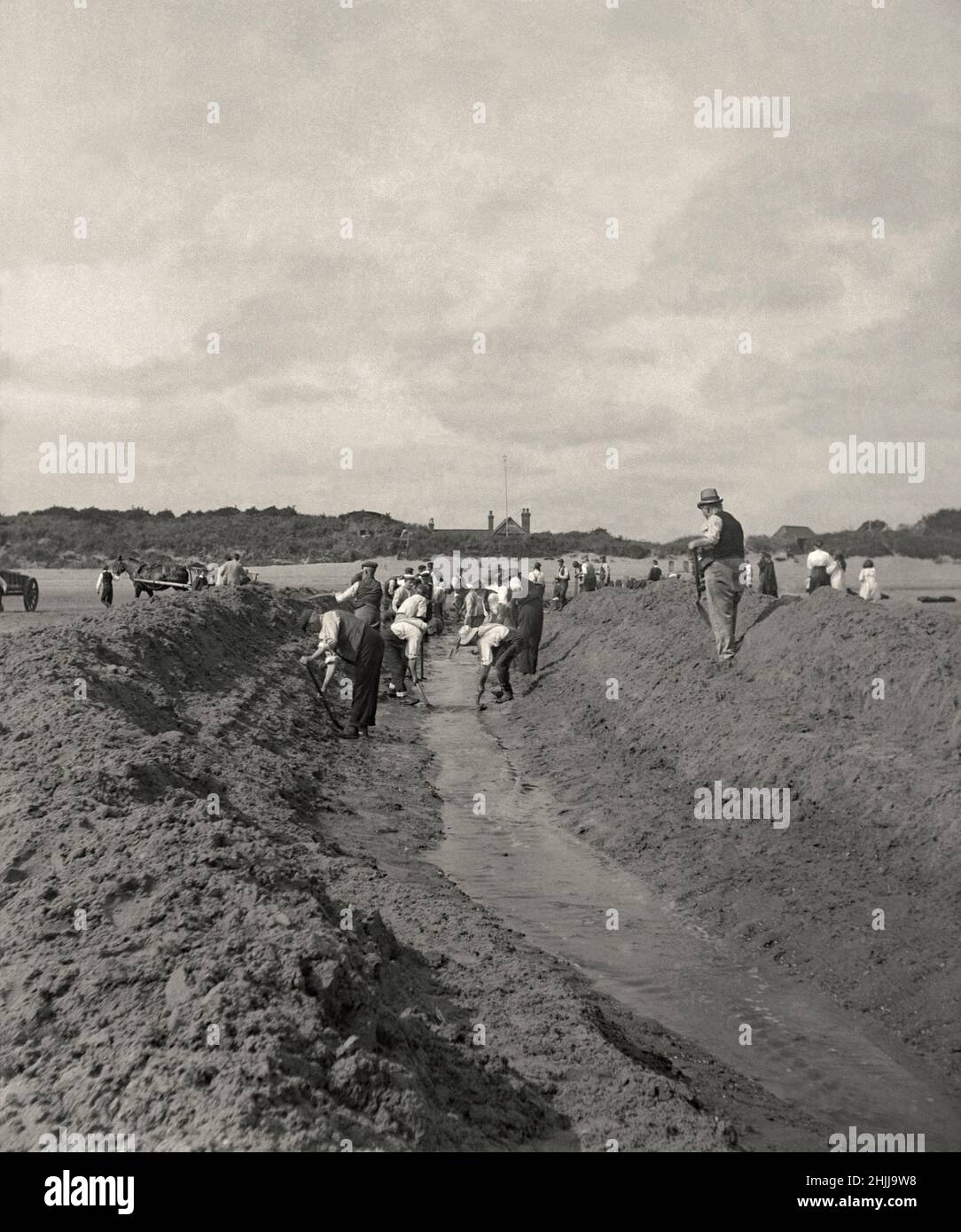 Digging or enlarging a drainage channel in the sand on a beach in Victorian Britain c. 1900. Sand is piled up on either side of the channel. A large team is digging by hand using spades and shovels. Horses and carts (left) are removing some of the spoil. The water seems to be flowing freely down the improved route. This is taken from an old black and white negative. It will look soft if used at too large a size – a vintage early sepia photograph. Stock Photo