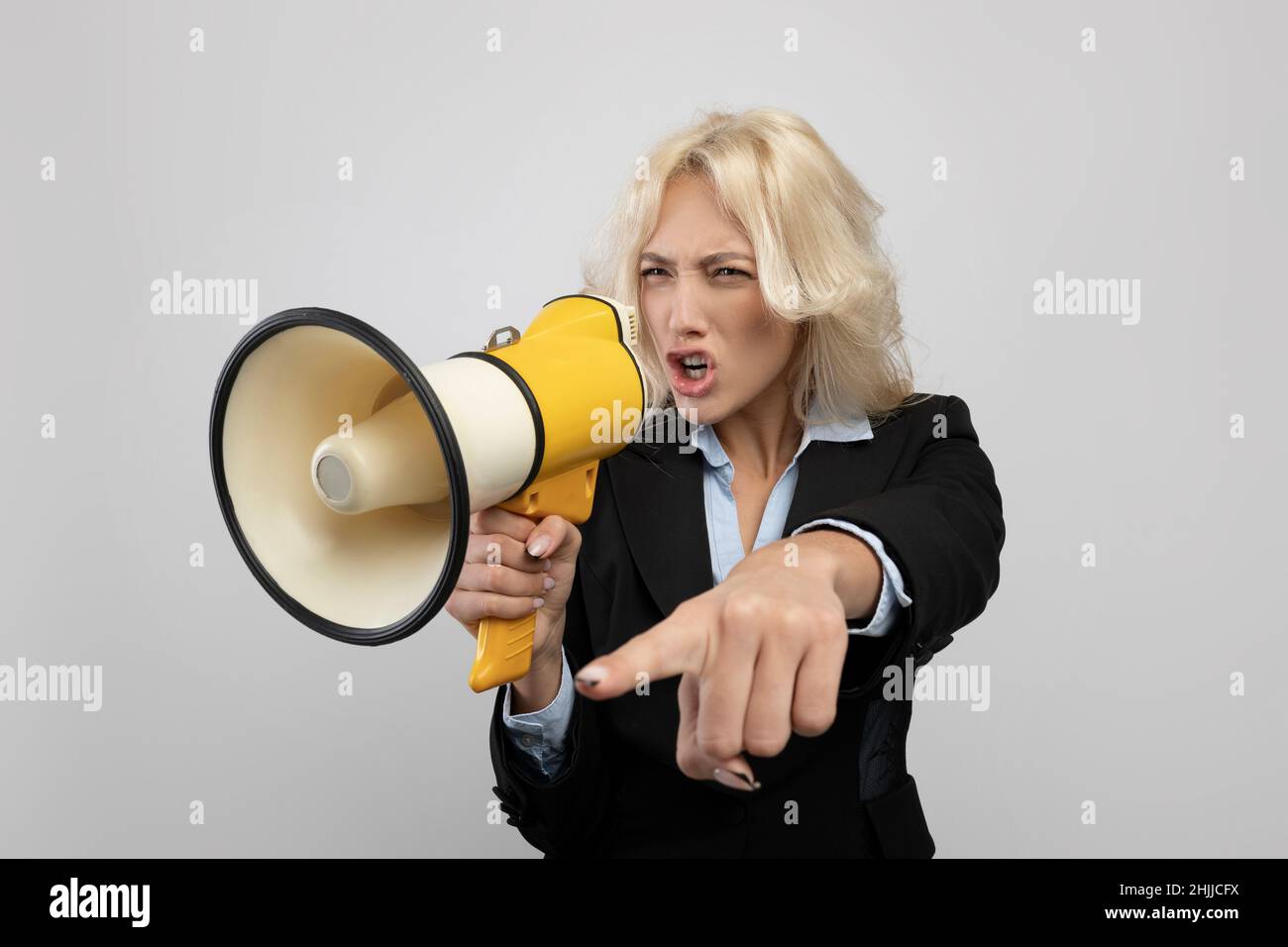 Agitated businesswoman in formalwear shouting into megaphone and pointing finger, posing on grey background Stock Photo