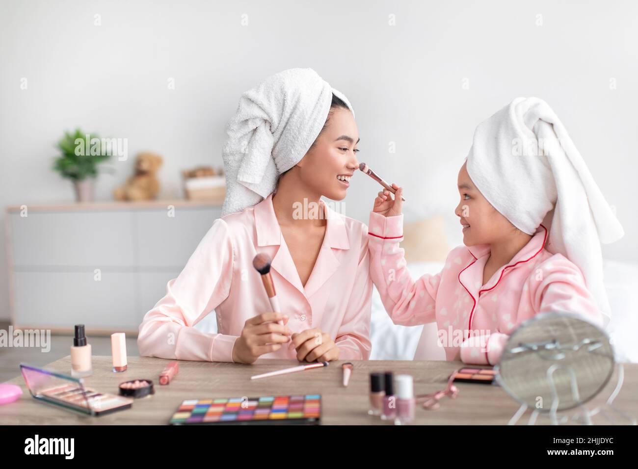 Cheerful chinese teenage girl and young female in pink pajamas and towels on heads do makeup in bedroom Stock Photo