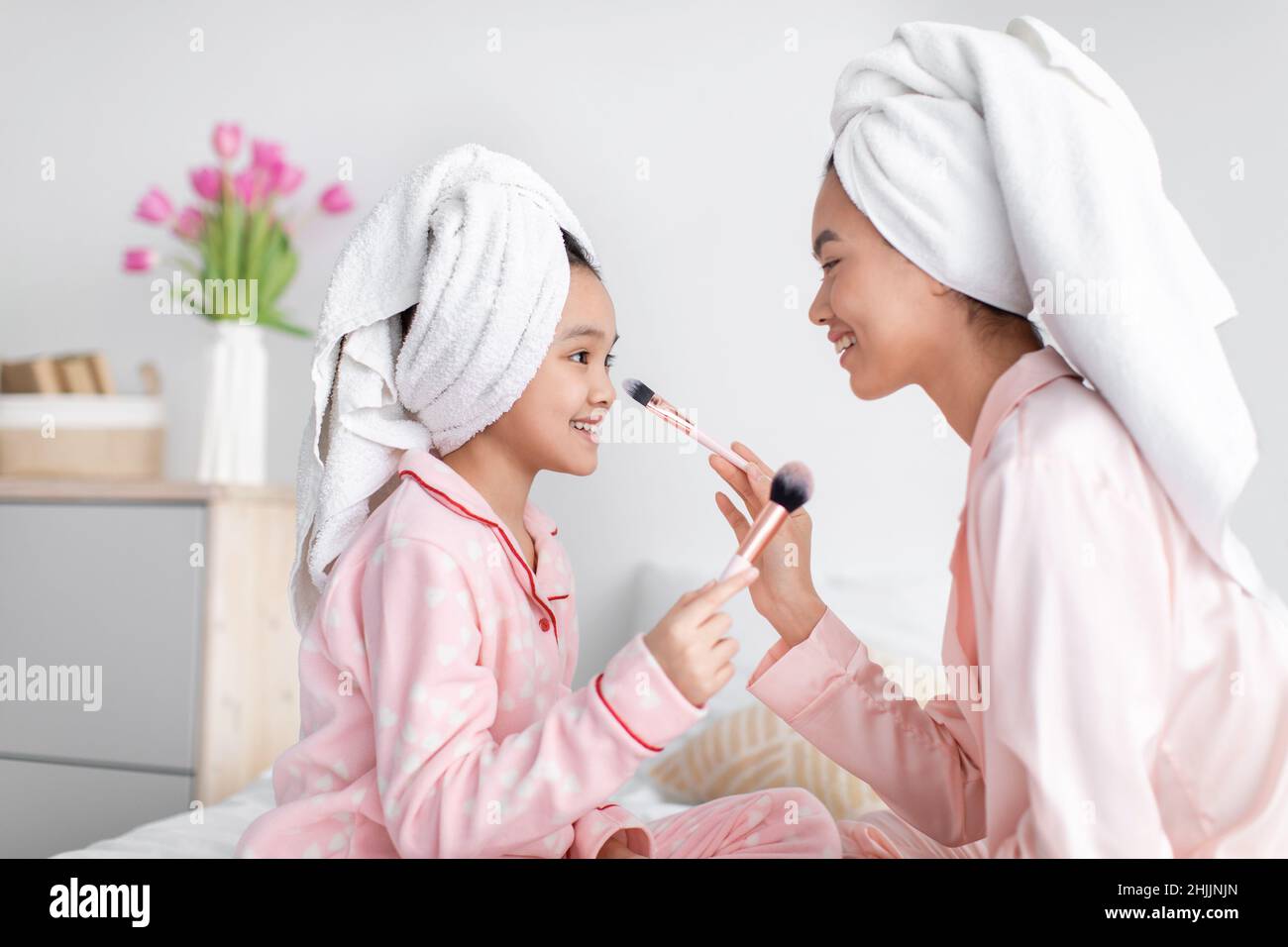 Happy cute korean teen girl and millennial female in pink pajamas and towel applying powder to face Stock Photo