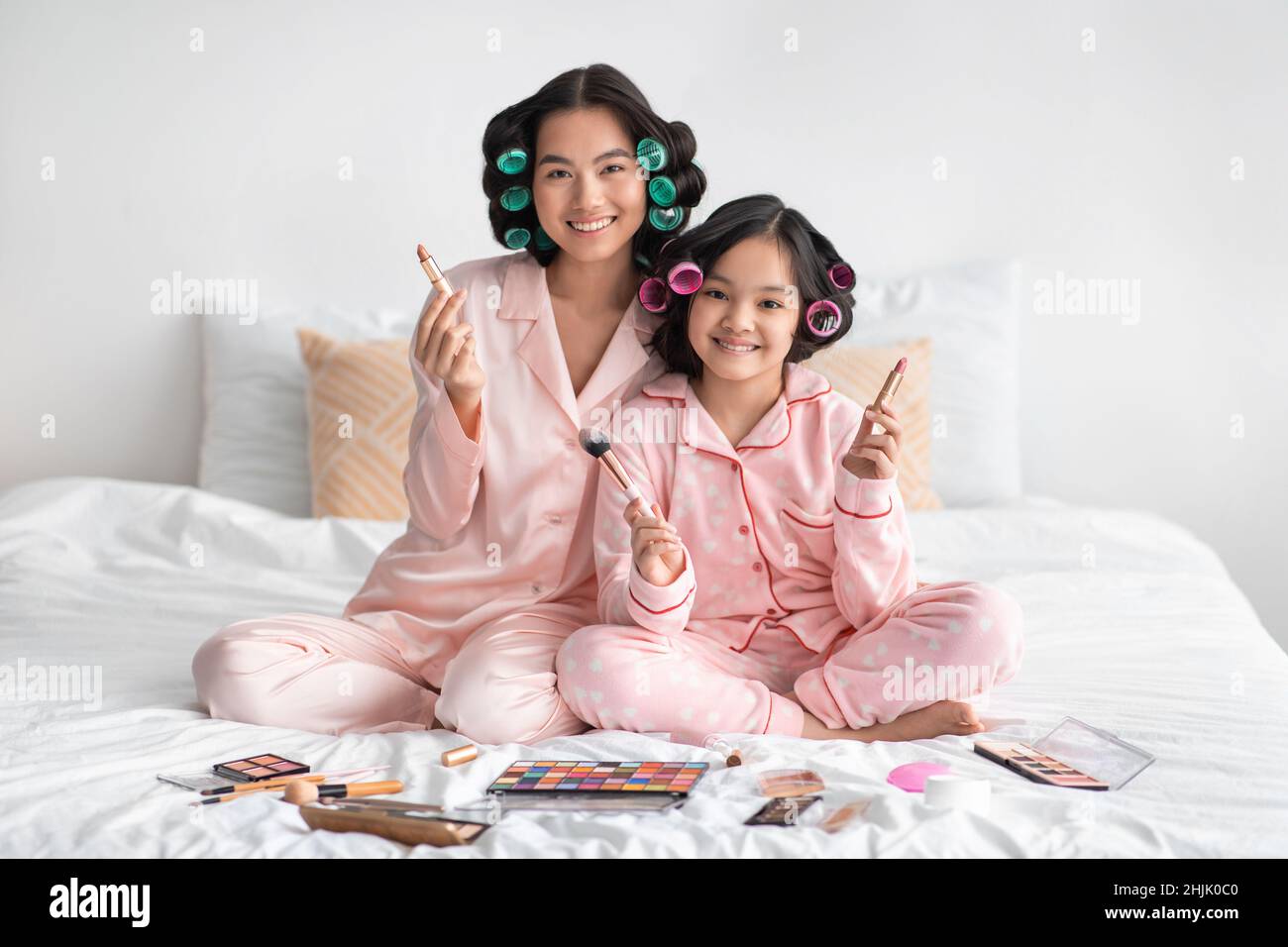 Happy millennial japanese female and adolescent girl with curlers hold brushes sit on bed with cosmetics Stock Photo