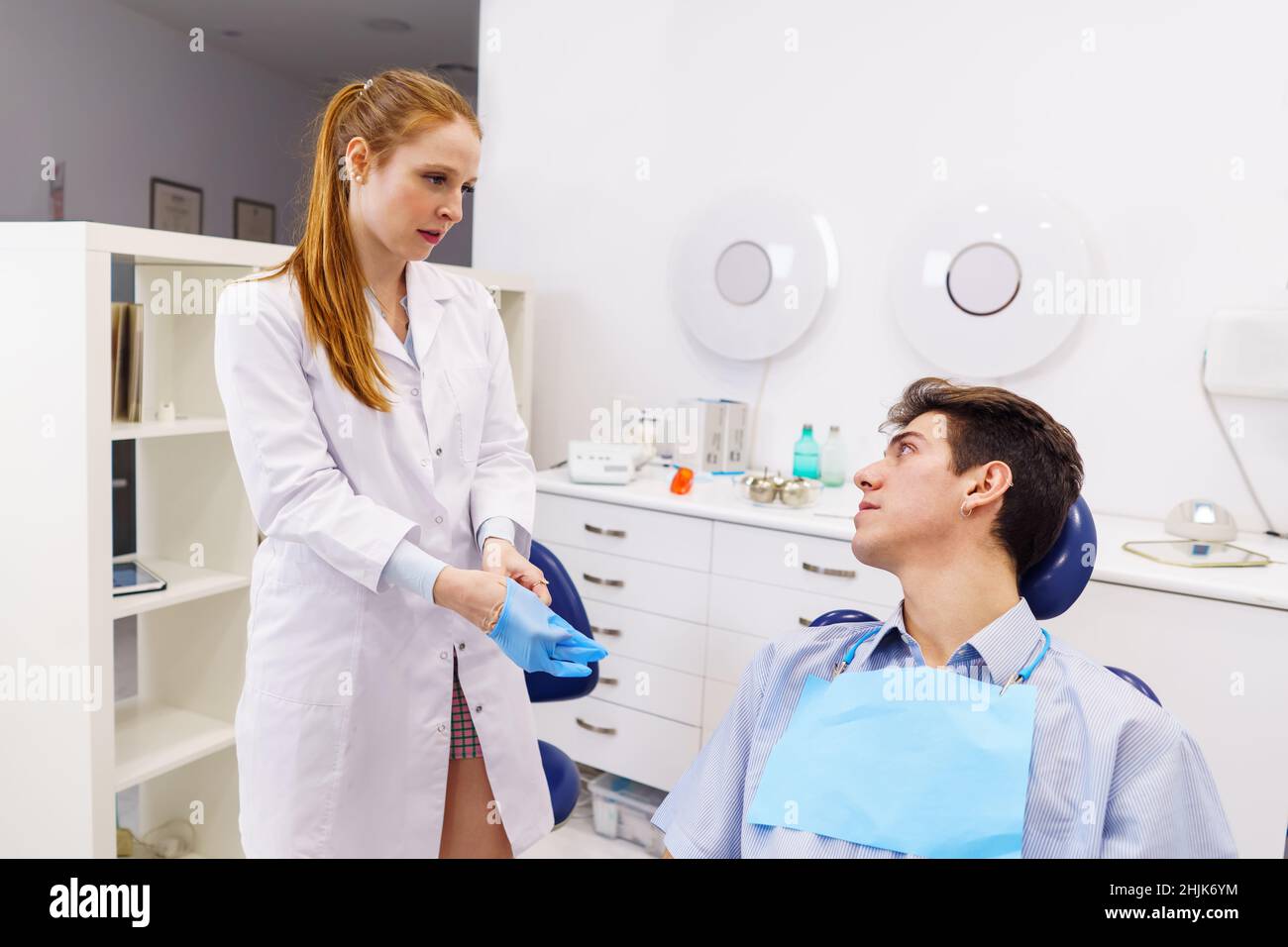 Ginger female doctor in white robe putting on latex glove and looking at man while working in