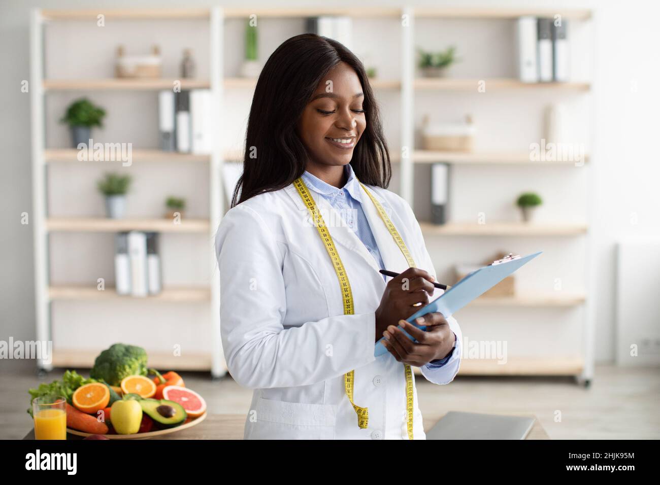 Cheerful black nutritionist with clipboard prescribing healthy diet or creating plant based meal plan for patient Stock Photo