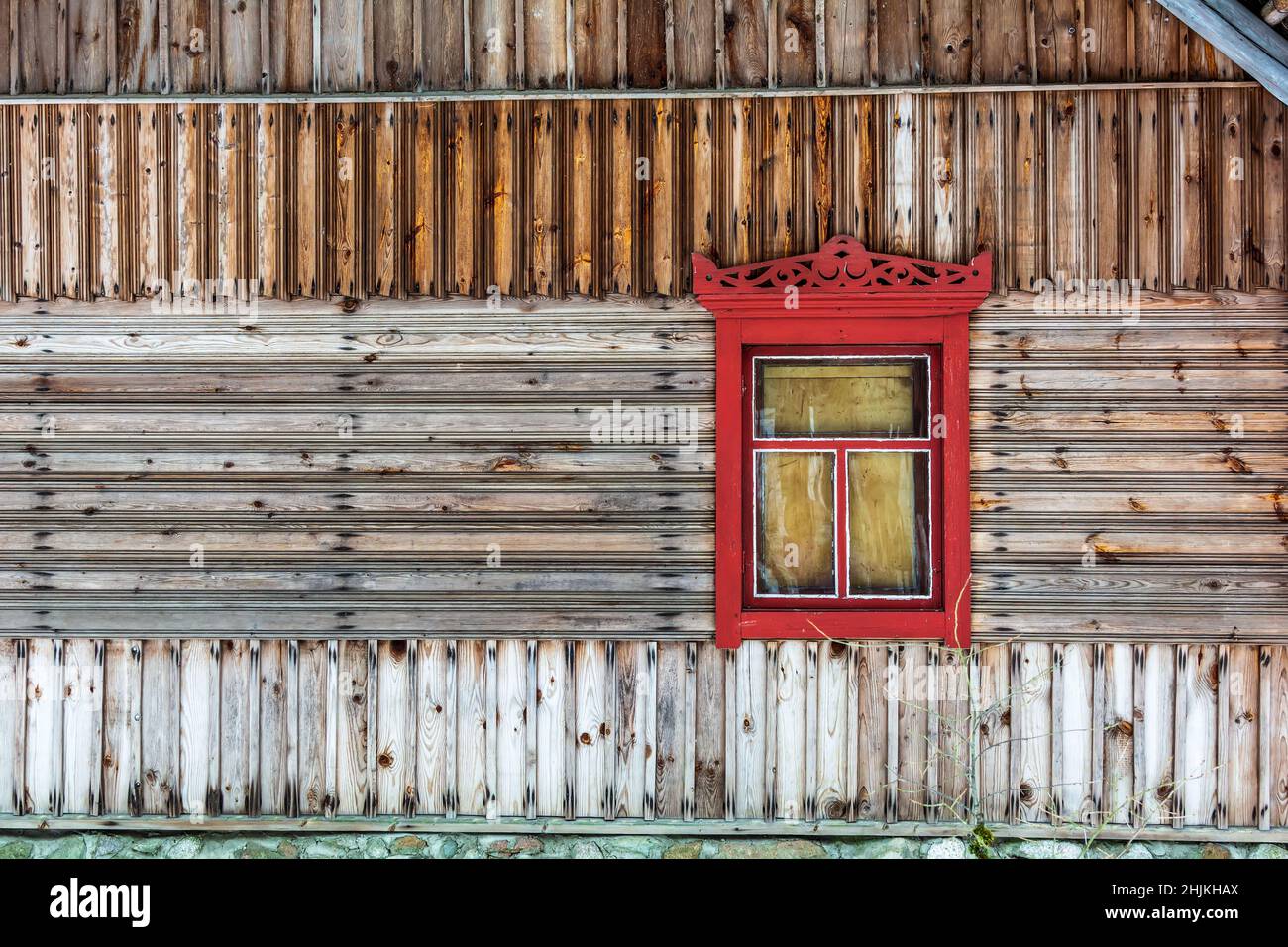 One vintage window with crimson red ornamental frame in brown wooden wall. Old dwelling-house of Russian peasant in Latvia. Stock Photo