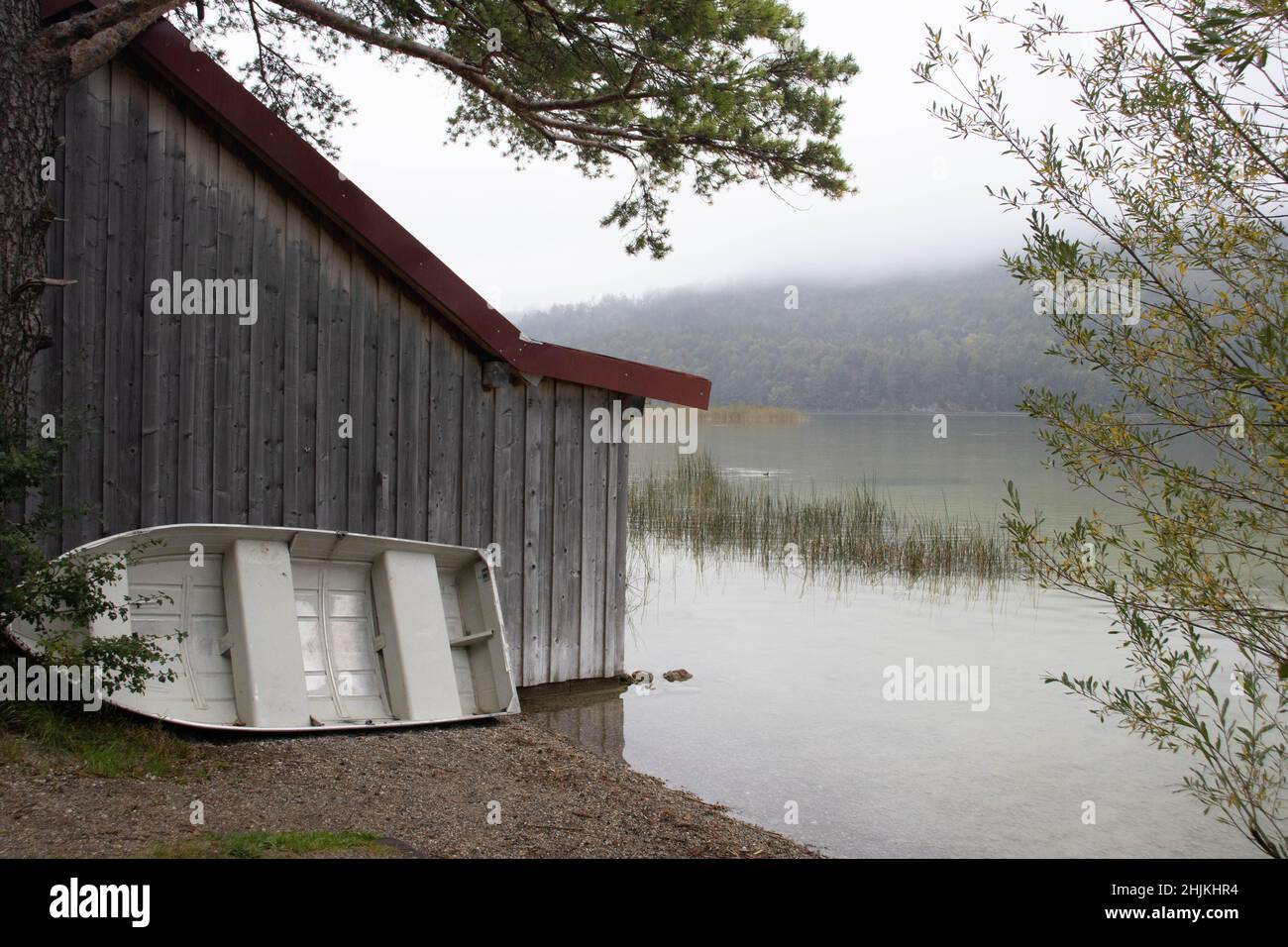 Ein weisses Anglerboot steht an einem Holzhaus (Bootshaus) am Ufer des Weissensees angelehnt Stock Photo