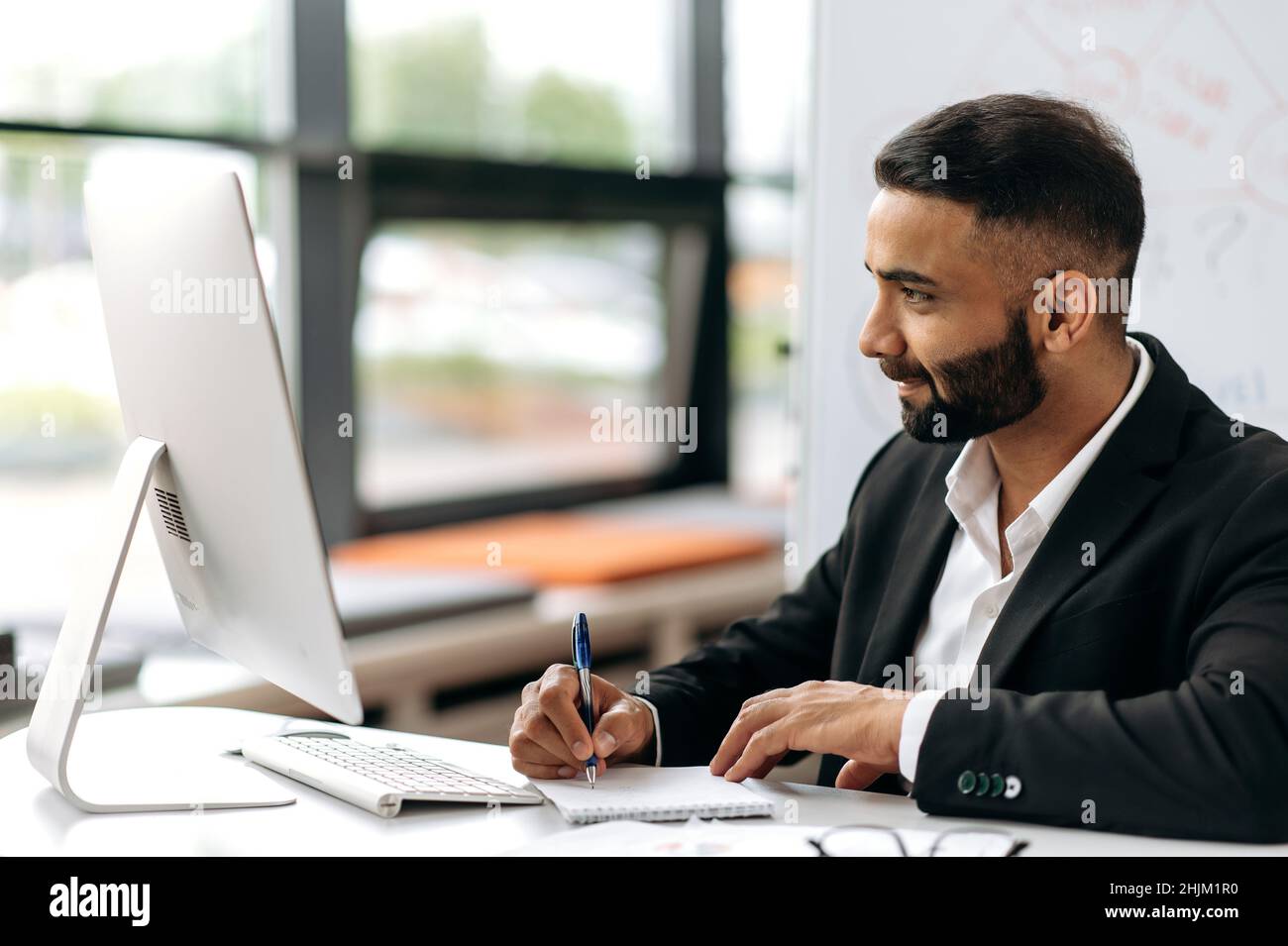 Smart attractive successful Indian businessman in suit, company director, financial advisor or broker sits at his desk, works on laptop, listening online conference, notes information, looks at screen Stock Photo