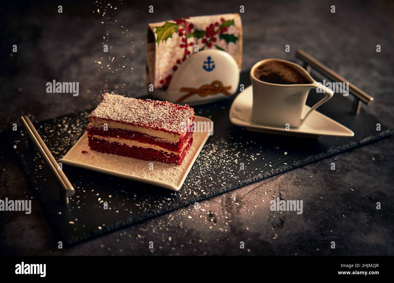 Sweet board.  Peas of cream cheese cake on a white ceramic plate with a cup of coffee and a napkin holder on a black ceramic tray. Stock Photo