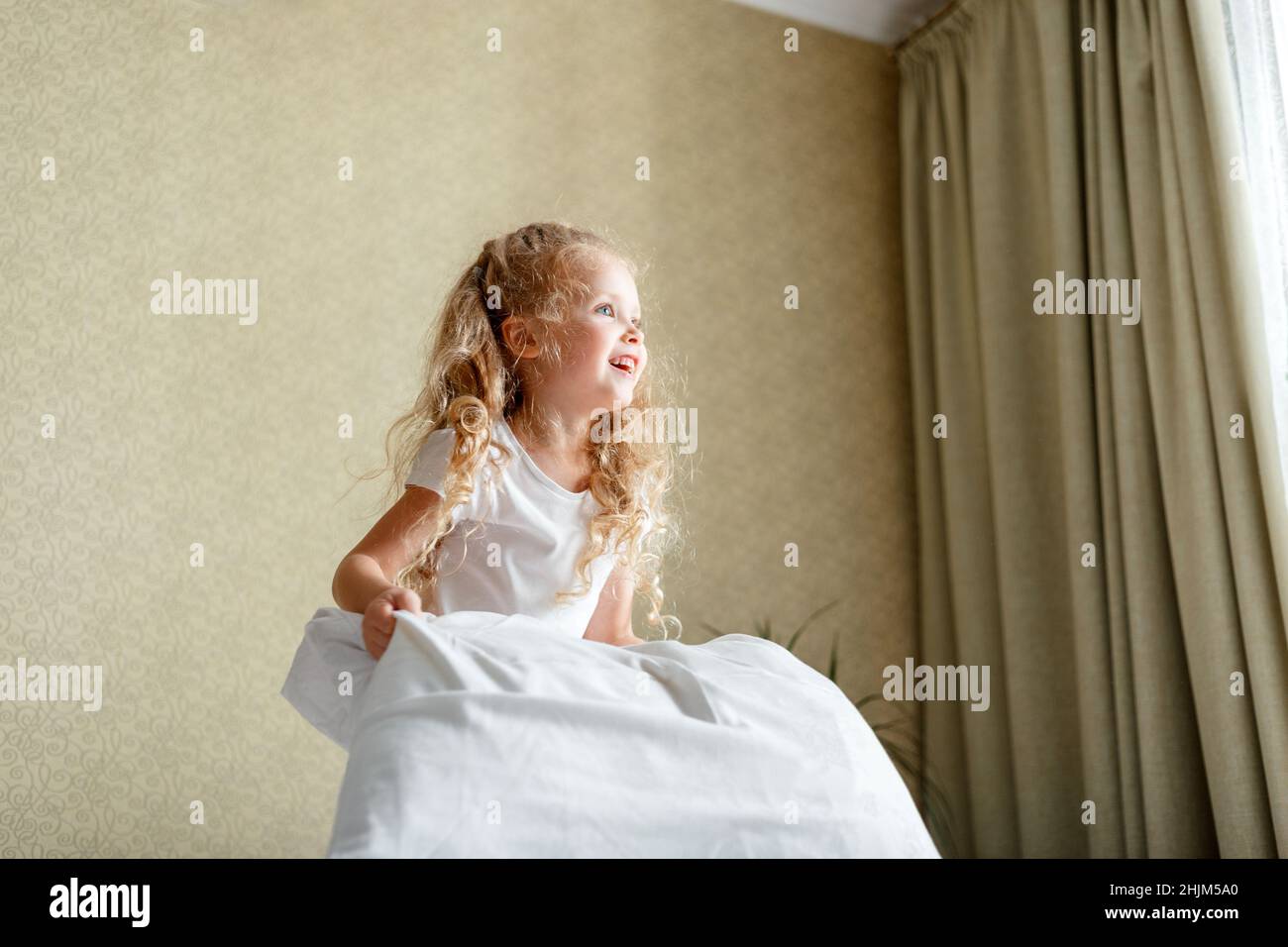 Cheery little Kid girl having fun jumping in bedoom playing with pillow. Portrait of preschool daughter Caucasian blonde girl play in pillows cushions Stock Photo