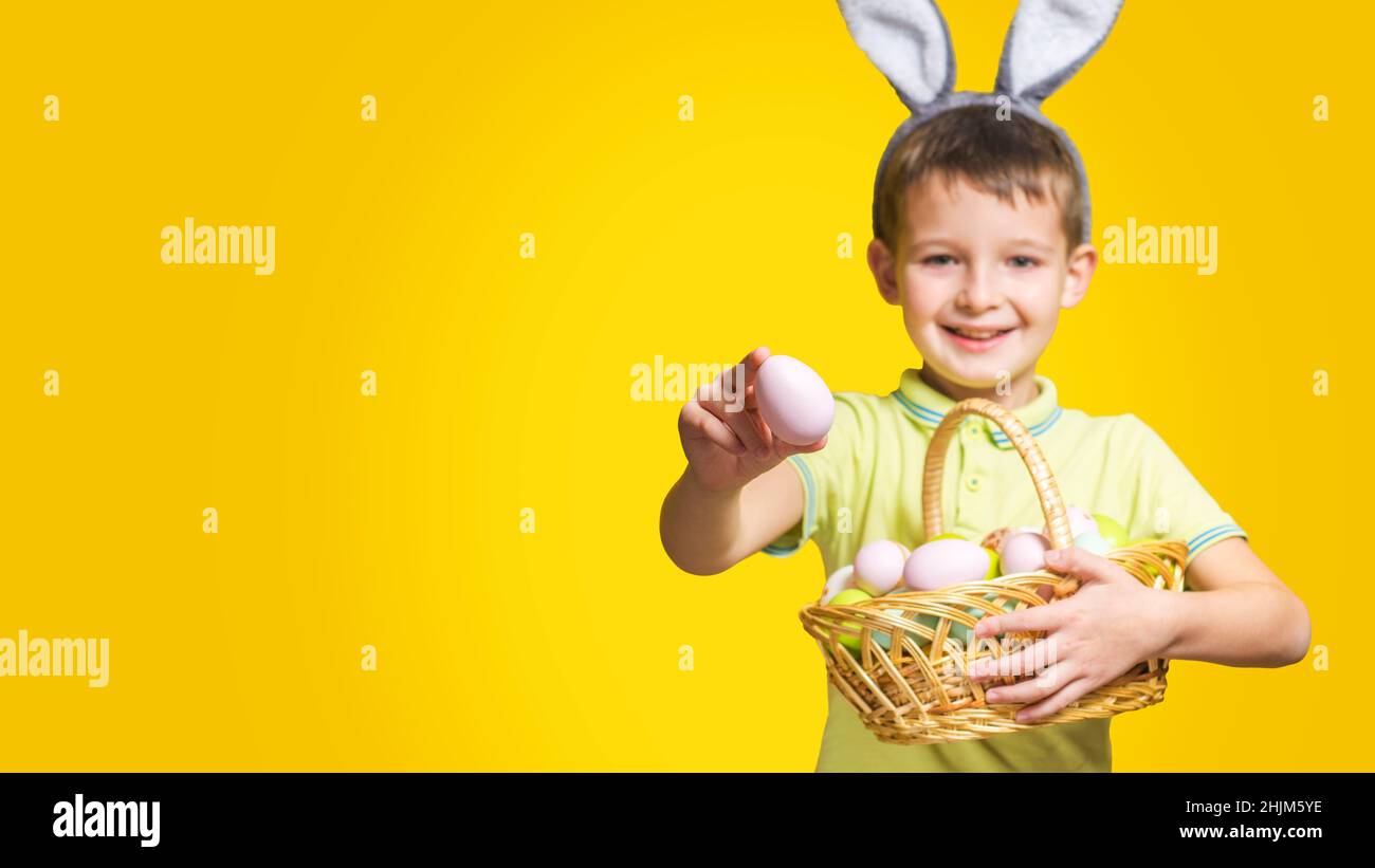 A happy smilling boy with Easter bunny ears holds a basket with painted eggs and shows one of them on a yellow background. Copy space. Easter day. Stock Photo