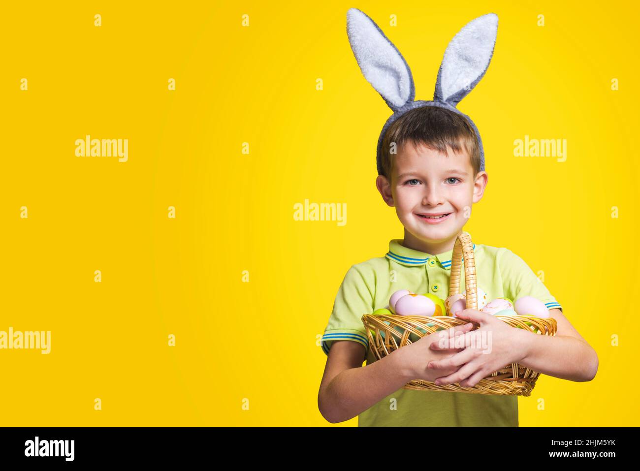 A happy smilling boy wearing Easter bunny ears holds a basket with painted eggs on a yellow background. Copy space. Easter day. Stock Photo