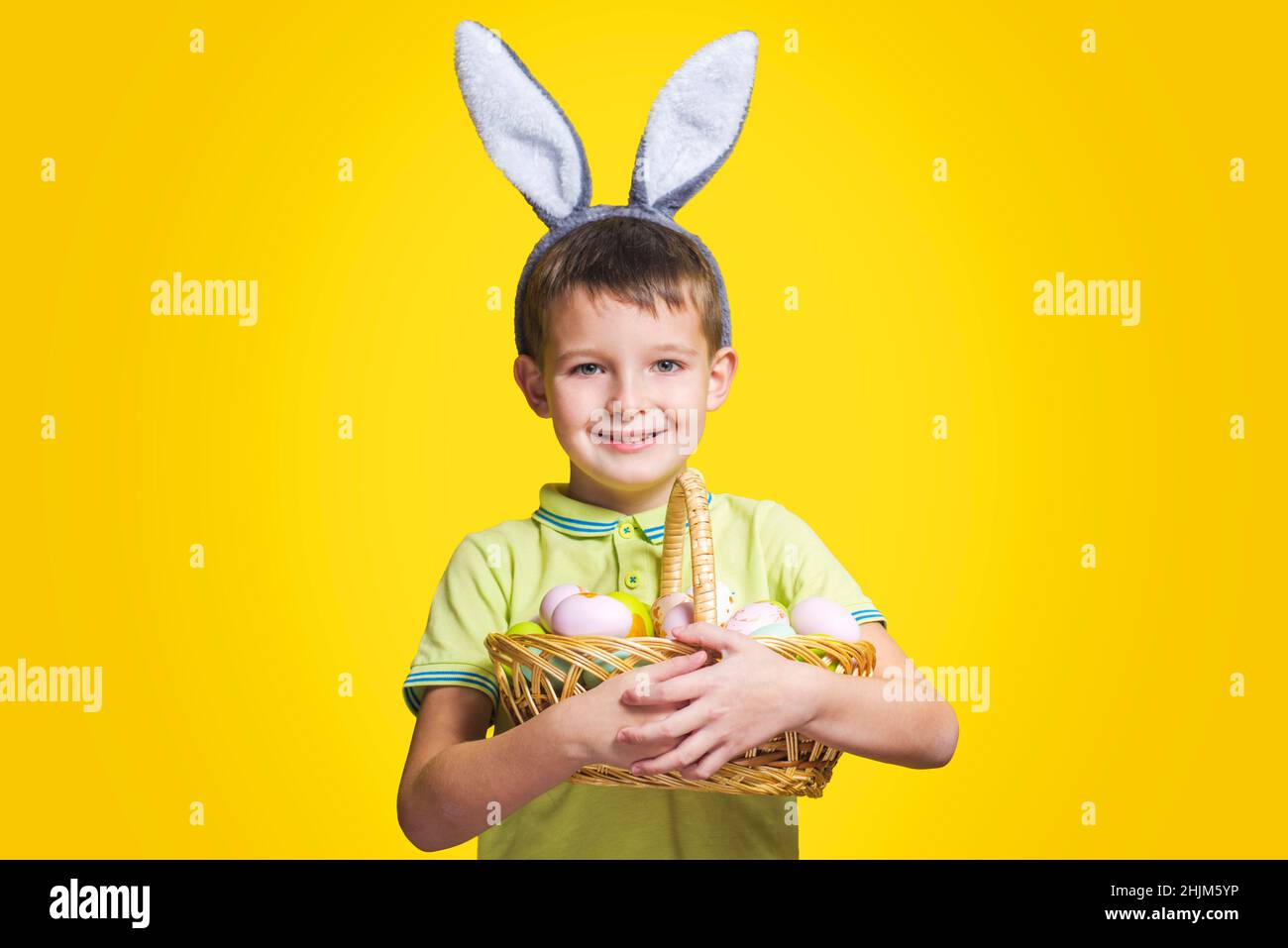 A happy smilling boy wearing Easter bunny ears holds a basket with painted eggs on a yellow background. Copy space. Easter day. Stock Photo