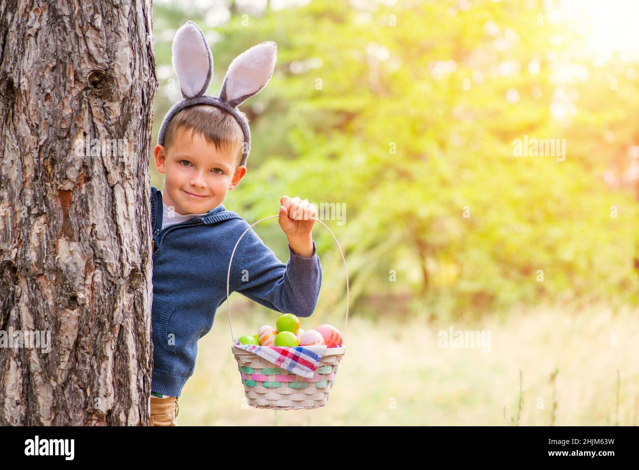 A cute little boy with a bunny ears standing behind a tree and holding basket full of colorful easter eggs after egg hunt in his hand. Happy Easter da Stock Photo