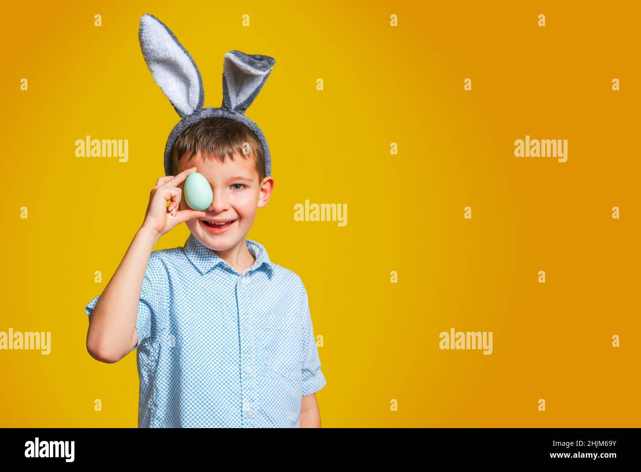 Happy child boy with bunny ears is covering his eye with Easter egg on yellow background. A kid with Easter egg on yellow background. Stock Photo