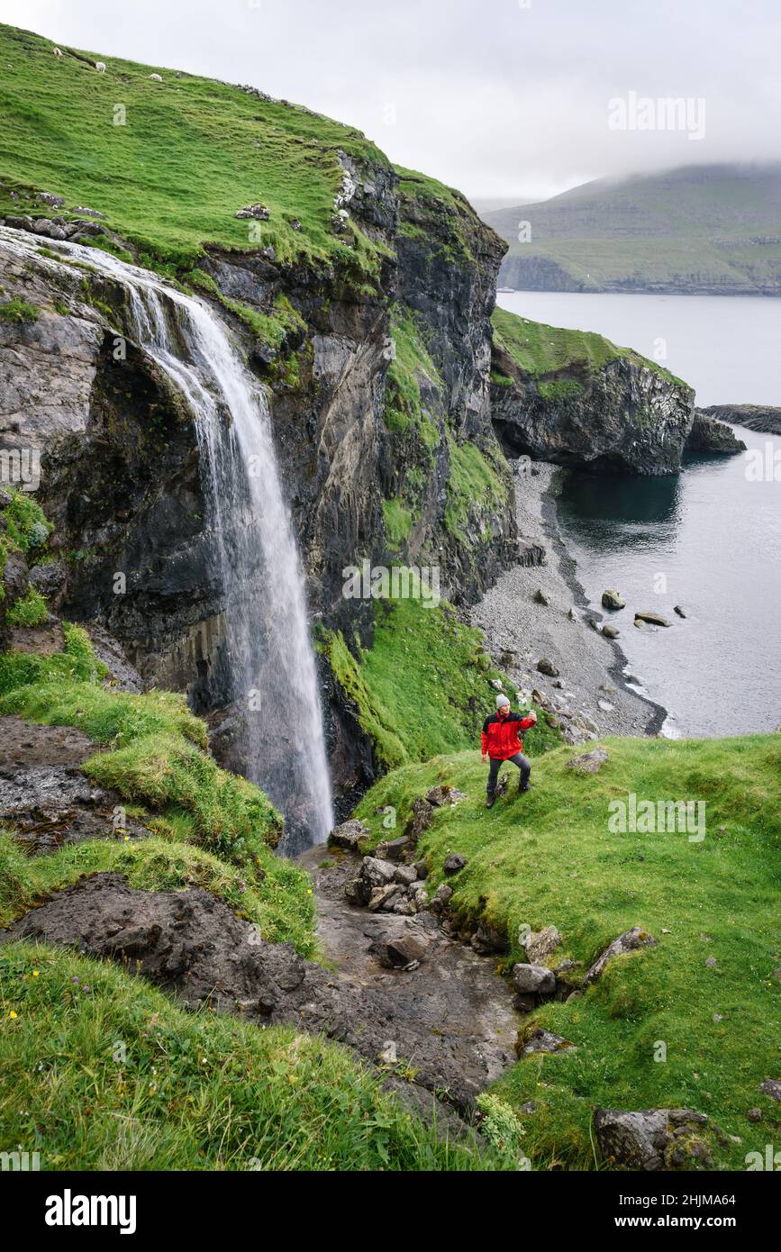 A traveler in a red jacket takes a selfie on the phone near the Skardsafossur waterfall on the island of Vagar, Faroe Islands Stock Photo