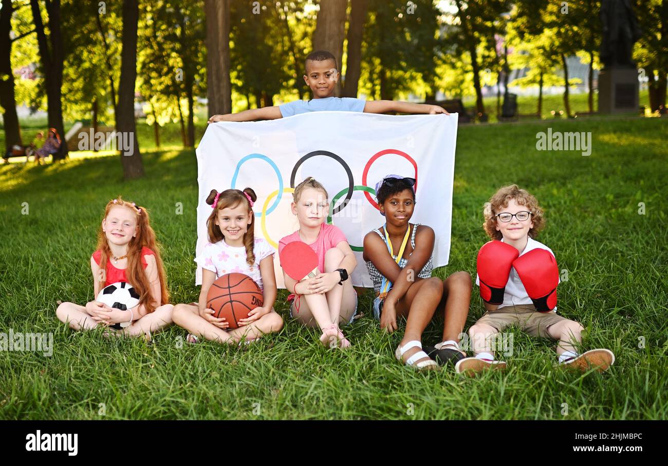 DONETSK, UKRAINE - JULY 10, 2021. Children of different nationalities with sports equipment in their hands hold a large flag with Olympic symbols Stock Photo
