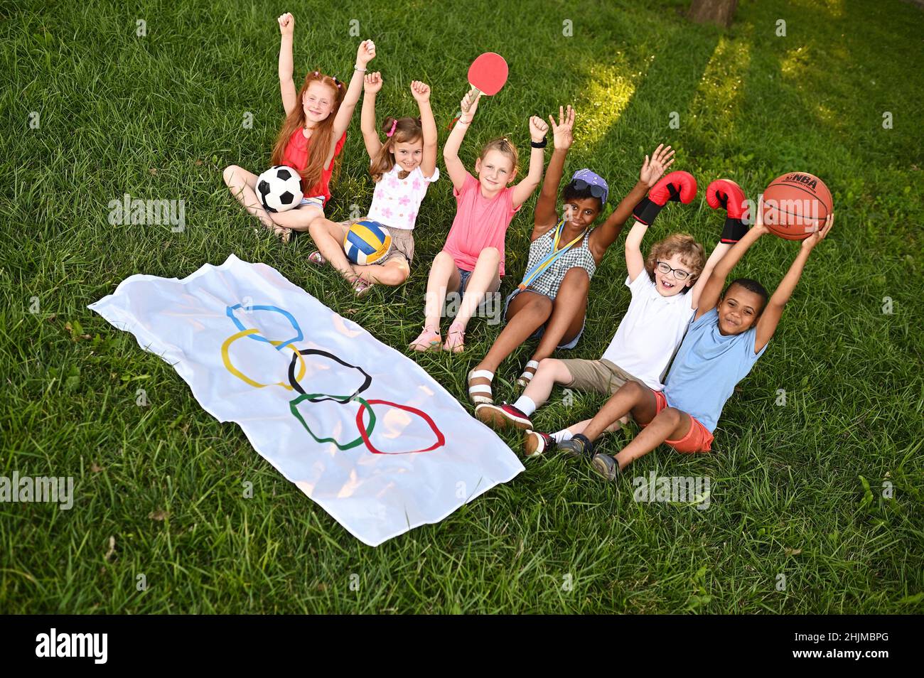 DONETSK, UKRAINE - JULY 10, 2021. Children of different nationalities with sports equipment in their hands are sitting on the grass against the Stock Photo