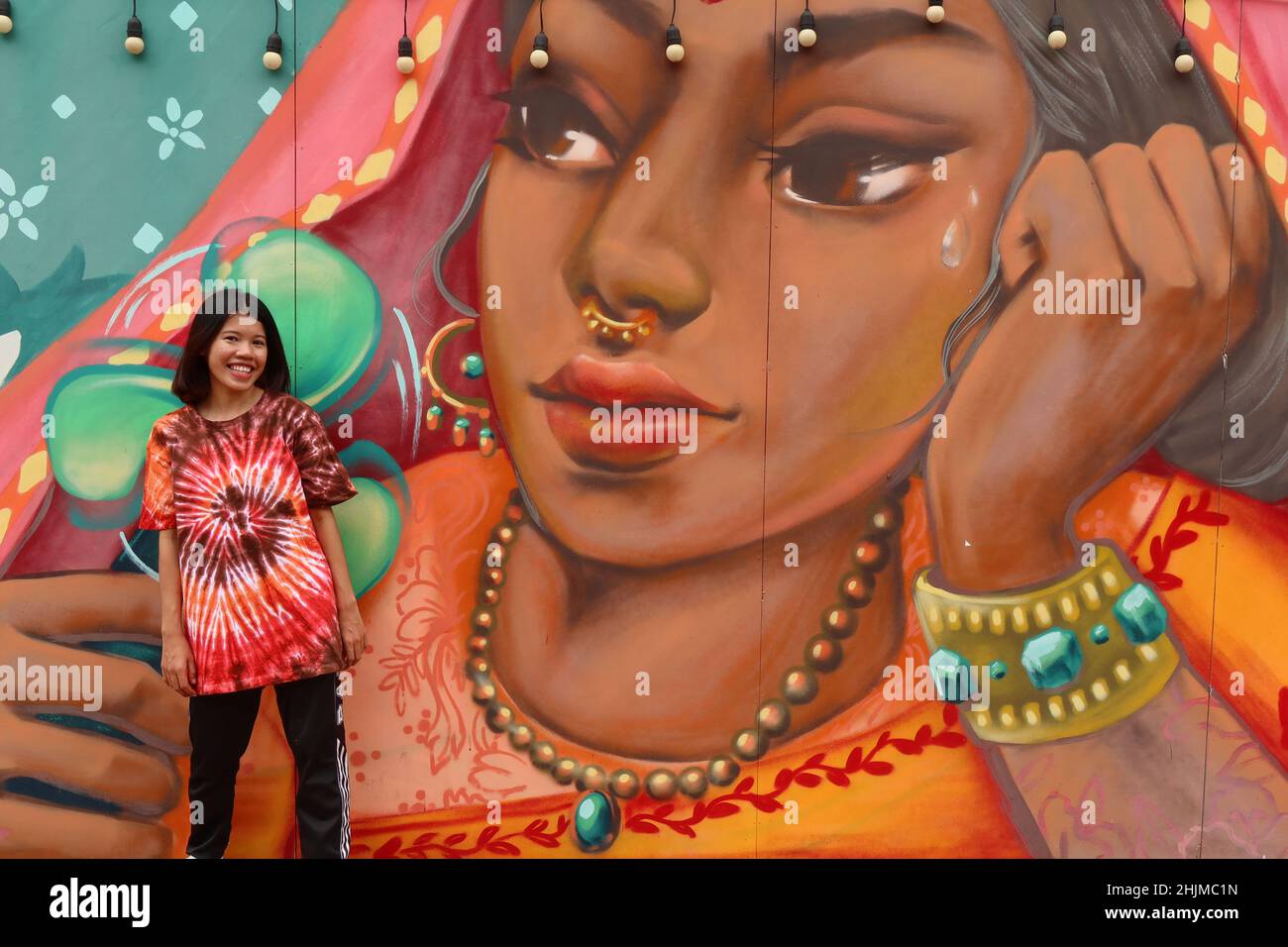 Woman smiling in front of a wall painting of an Indian woman on Ong Ang Walking Street by Klong Ong Ang, Chinatown, Phahurat, Bangkok, Thailand, Stock Photo