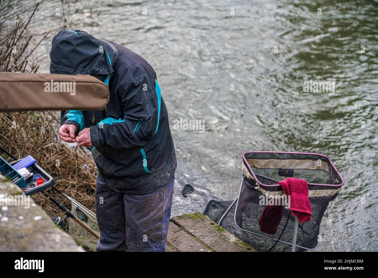 fish angler stood on a wooden jetty prepares his fishing hook bait to cast in to the flowing river avon, Salisbury UK Stock Photo