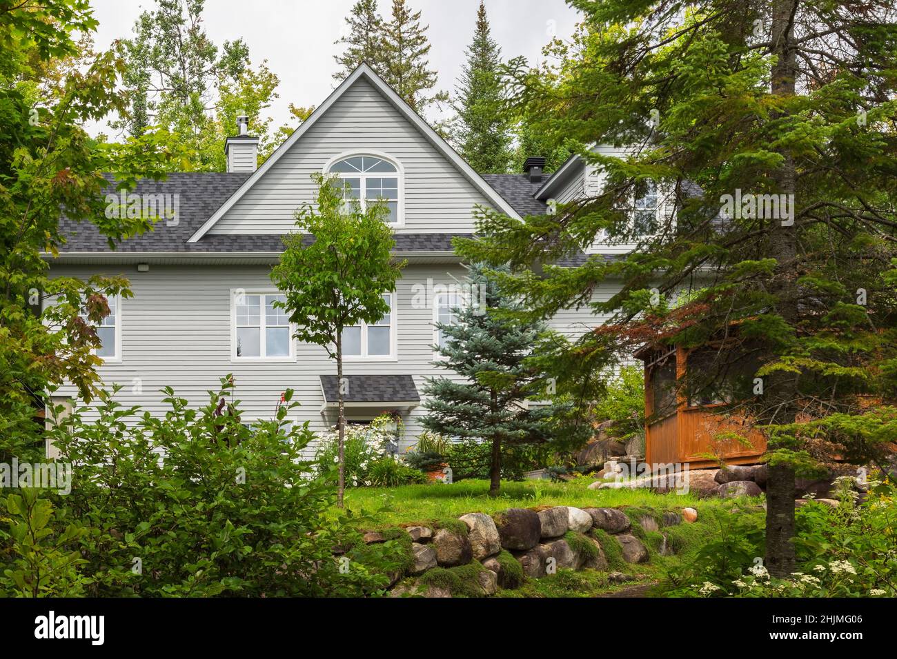 Back of cottage style house through deciduous and conifer trees with raised terrain bordered by rock wall with Thymus serpyllum - Wild Thyme. Stock Photo