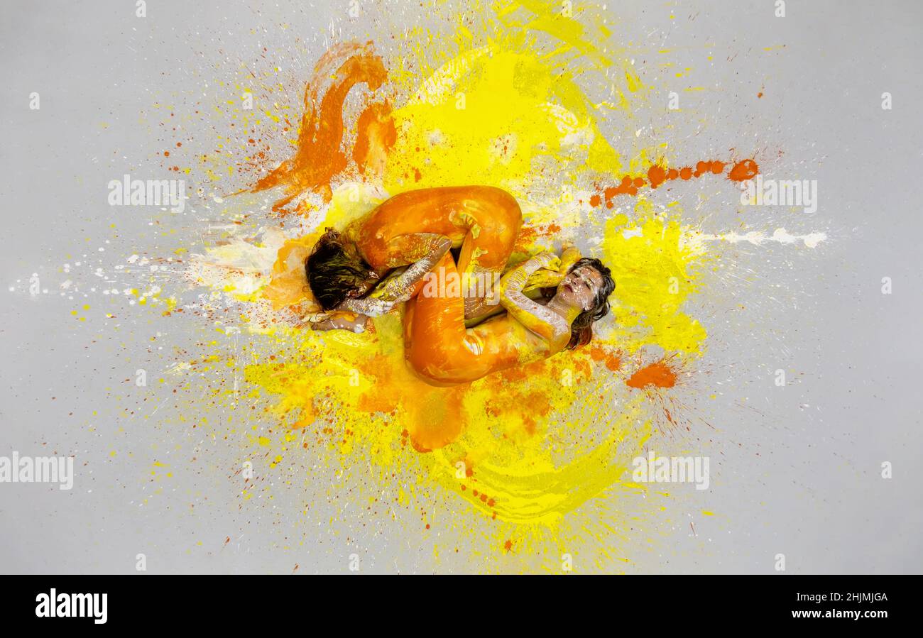 two yellow and orange abstractly painted young women lying huddled on the artists studio floor in fetal position, wildly speckled with colorful paint Stock Photo
