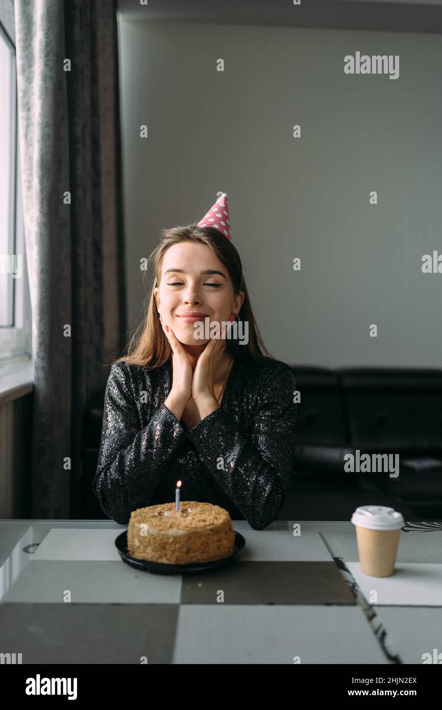 Happy caucasian young woman celebrating birthday at home, girl sitting at table has cake with candle, holiday at home in quarantine, birthday Stock Photo