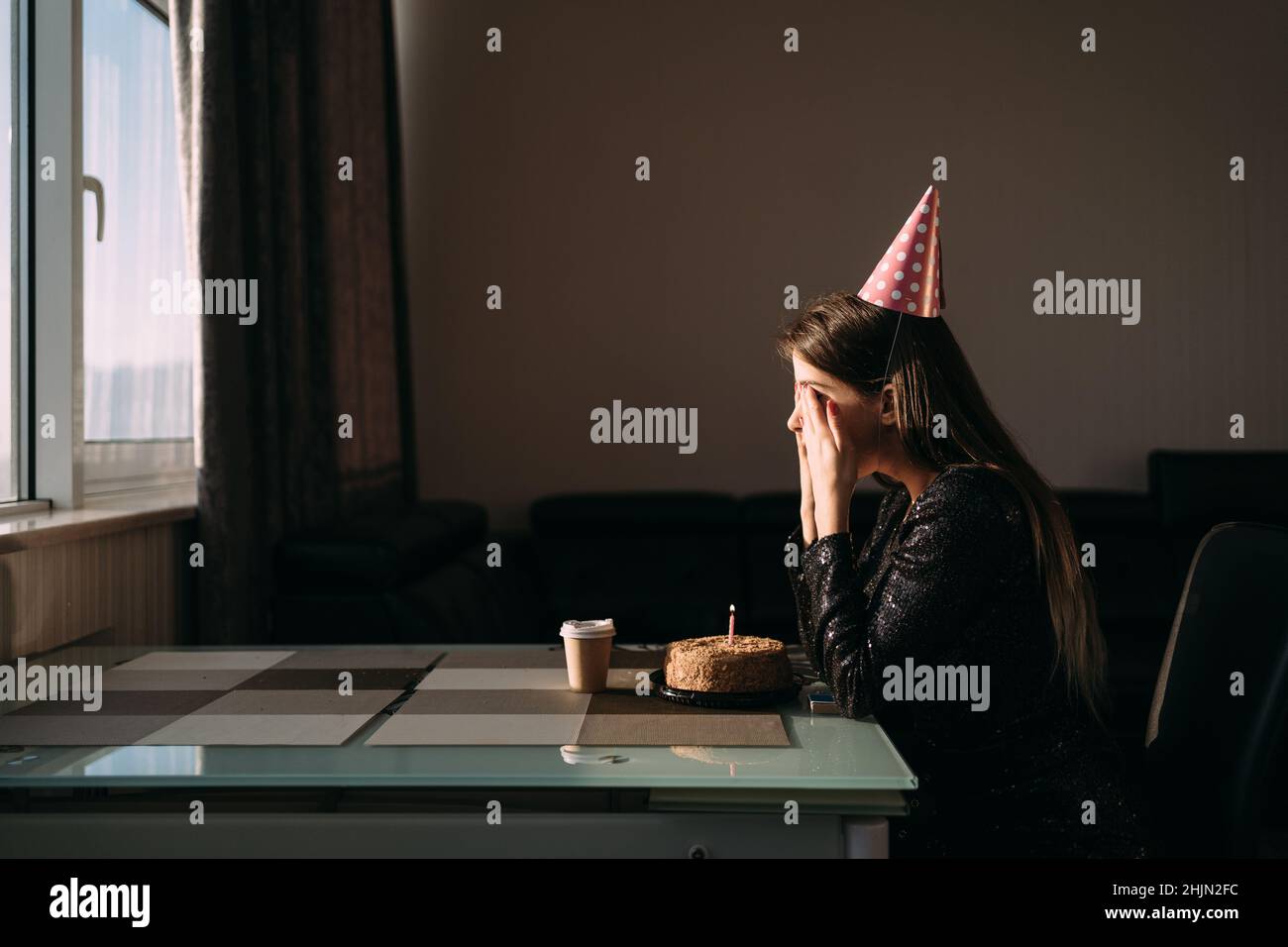 Cute thoughtful woman in party hat looking at festive cake with candle. Lonely woman freelancer celebrating birthday without friends. Stock Photo