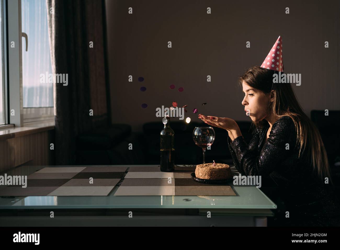 isolated birthday at home, woman in festive hat alone at the table, lonely holiday freelancer, cake and confetti, festive background, introversion Stock Photo
