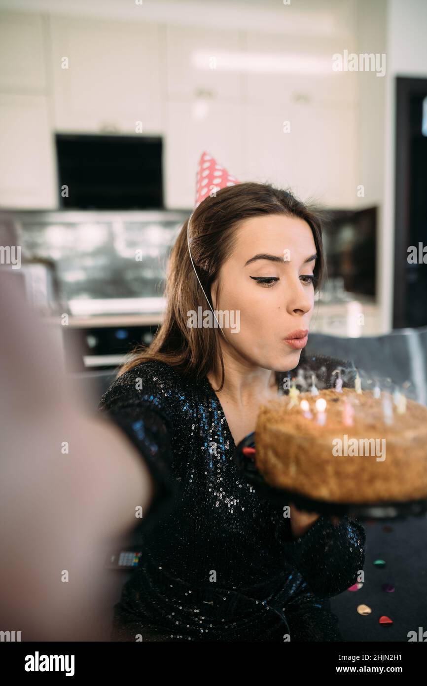 Birthday online, woman blows out the candles on the cake, girl in a festive cap at home with cake, video chat with friends, blogger anniversary on Stock Photo
