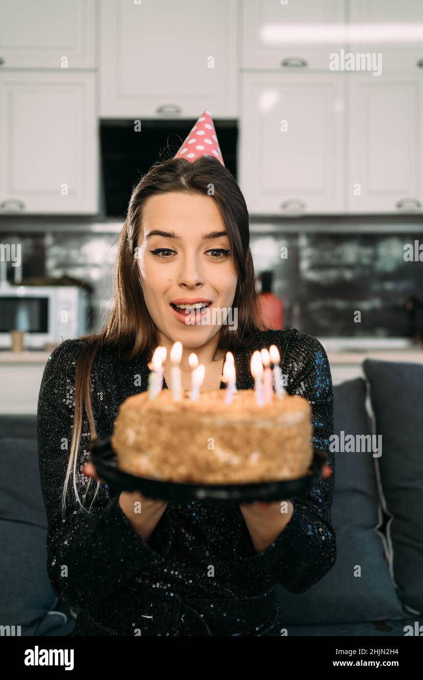 Young woman blowing out candles on a birthday cake, a girl alone in a party hat blows a candle, celebration at home, happy birthday concept Stock Photo