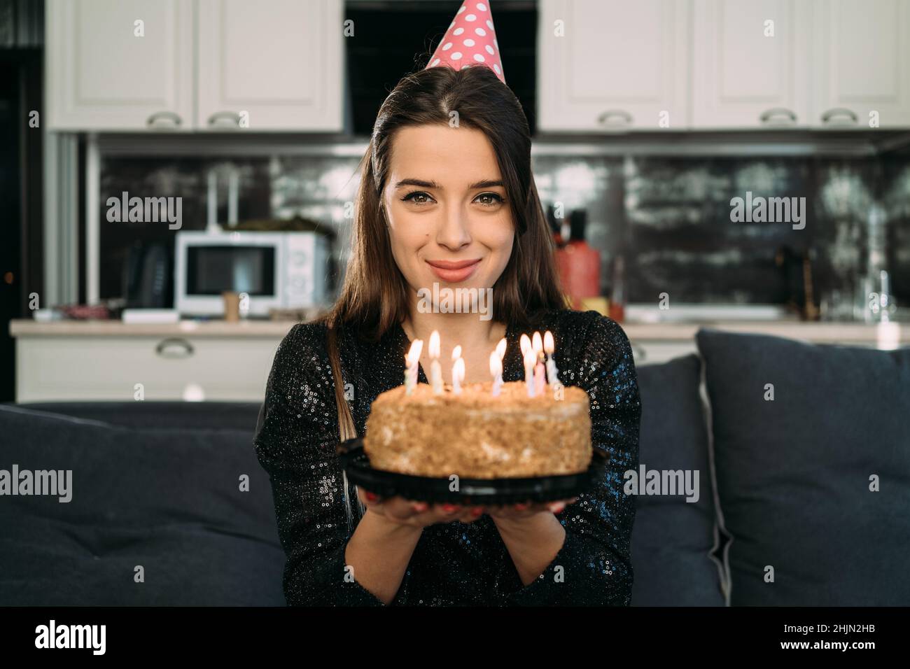 Portrait of a caucasian brunette with a birthday cake with candles, a girl in a cap at home alone celebrates an anniversary Stock Photo