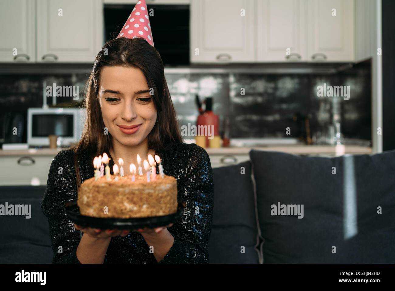 A young Caucasian lady is sitting at home on the sofa on her head wearing a birthday cap and holding a birthday cake with candles Stock Photo