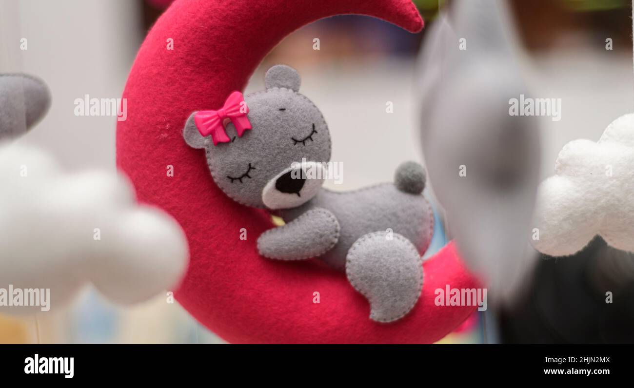 Baby crib mobile ornaments hangings close up shot, grey wool felt bear sleeps on a red plushie moon, surrounded by soft clouds. Stock Photo