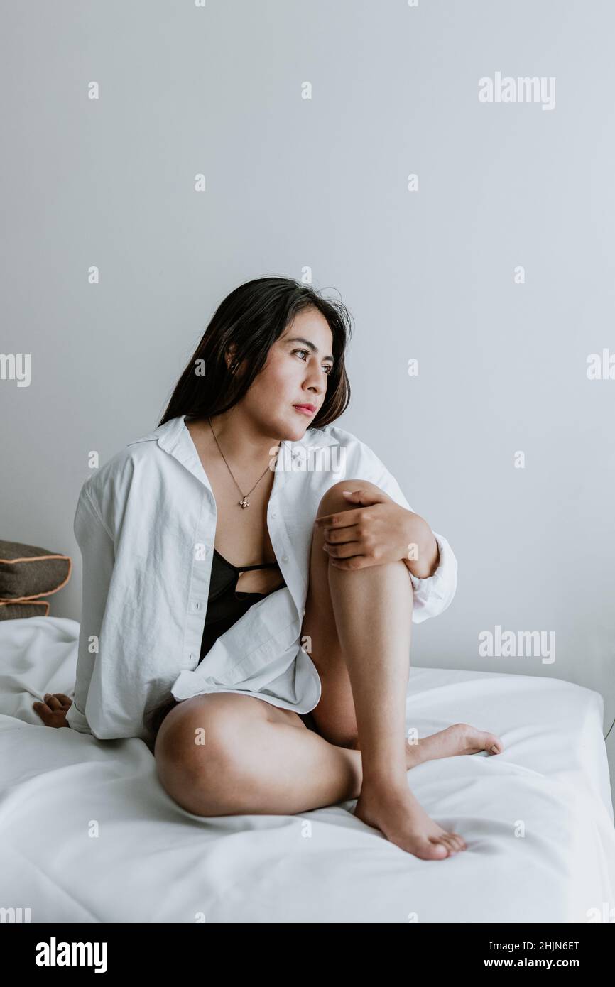 Young Latin woman sitting on bed, wearing white robe, showing legs at home in Mexico city Stock Photo
