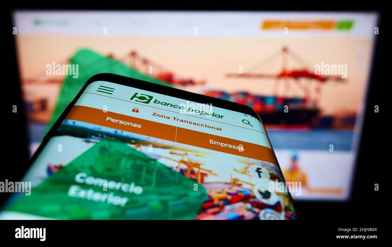Mobile phone with webpage of Colombian banking company Banco Popular S.A. on screen in front of monitor. Focus on top-left of phone display. Stock Photo
