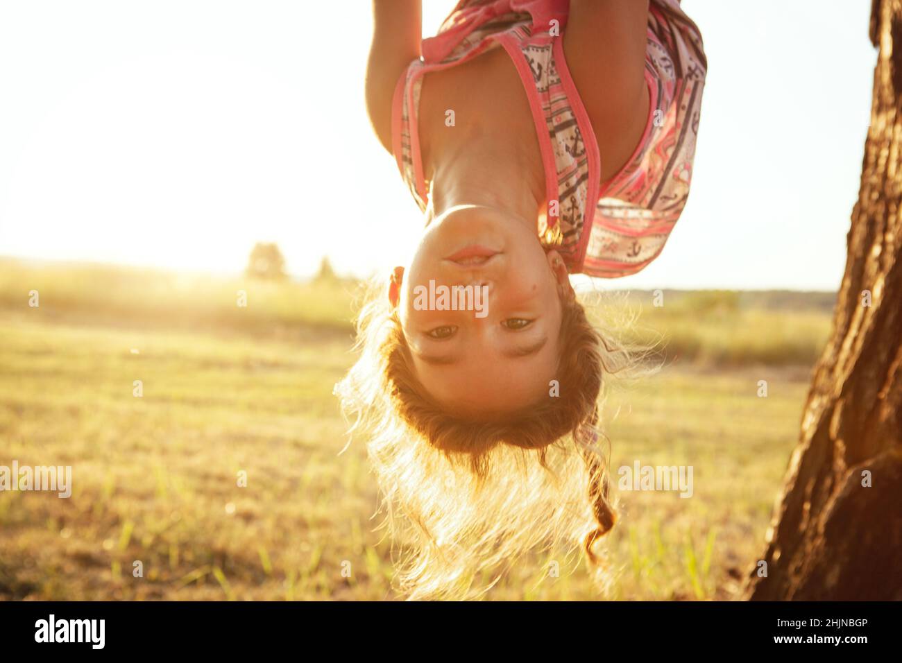 The girl is hanging upside down on a tree in summer in orange sunlight and a light dress. Summer time, heat, childhood. Funny portrait with disheveled Stock Photo