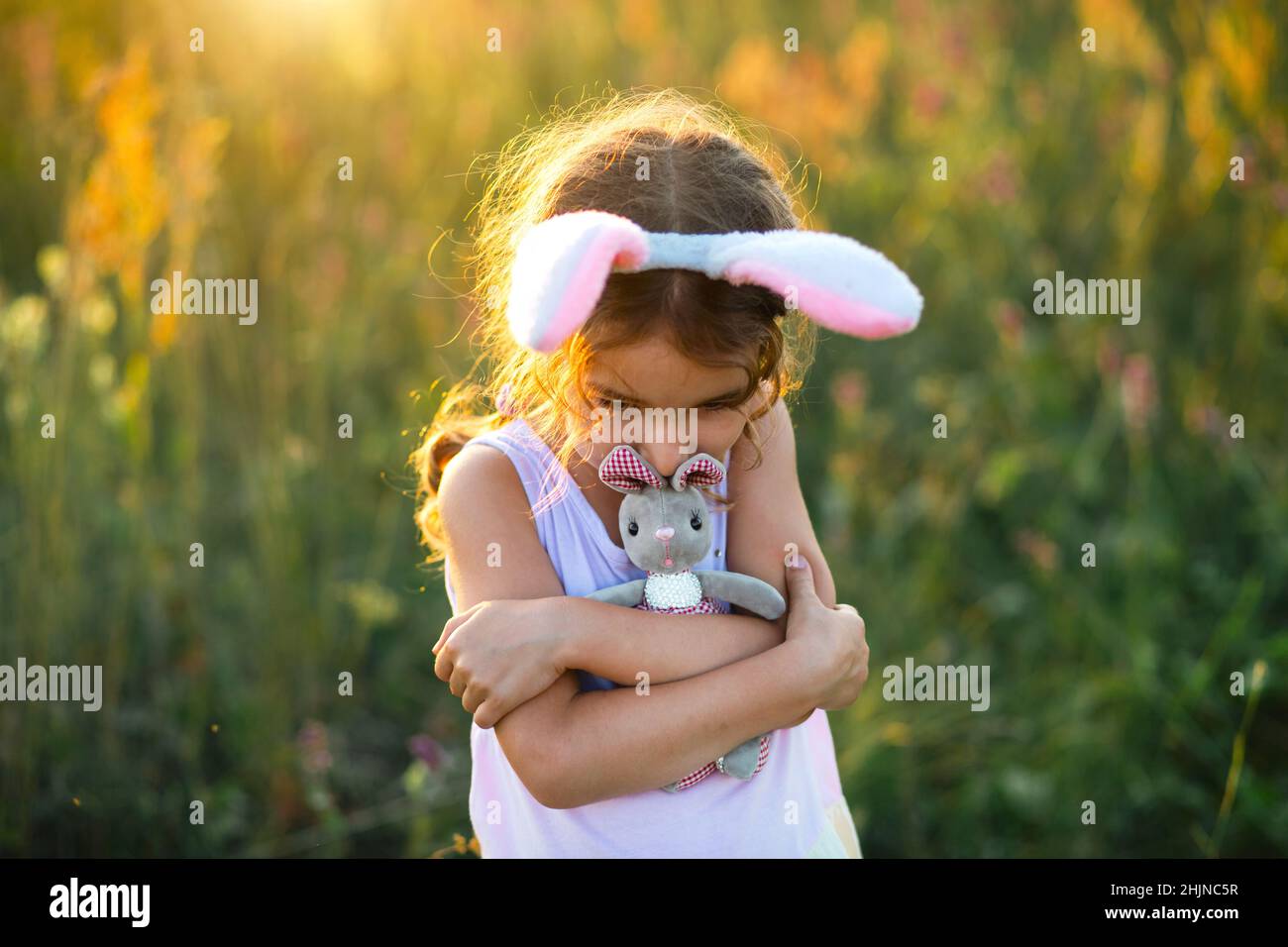 Cute 5-year-old girl with rabbit ears gently hugs a toy rabbit in nature in a blooming field in summer with golden sunlight. Easter, Easter bunny, chi Stock Photo