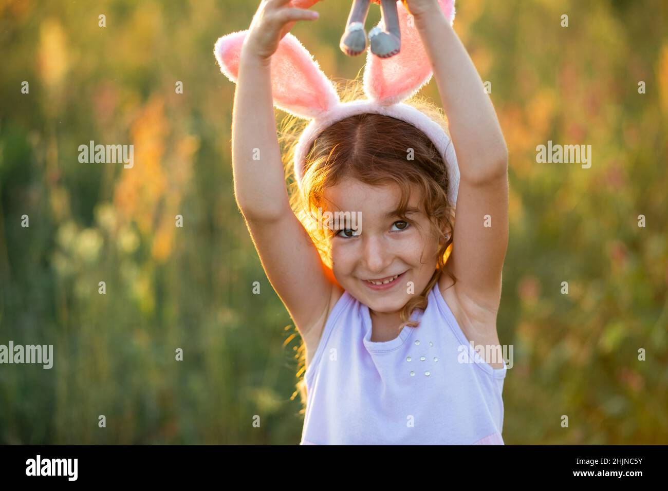 Cute 5-year-old girl with rabbit ears gently hugs a toy rabbit in nature in a blooming field in summer with golden sunlight. Easter, Easter bunny, chi Stock Photo