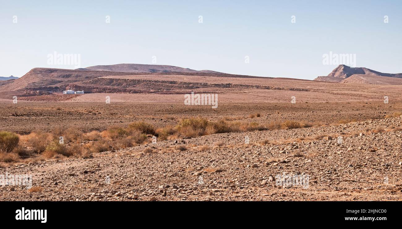 panorama of the bottom of the Maktesh Ramon crater showing one solitary campsite with sparse vegetation in the foreground and a clear blue sky in the Stock Photo