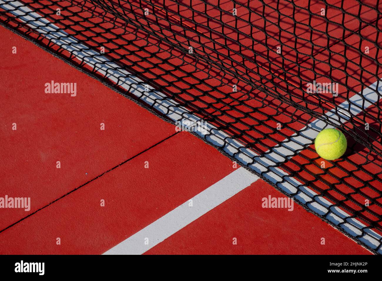 Tennis ball next to the net of a red hard surface tennis court. Racket sports concept. Stock Photo