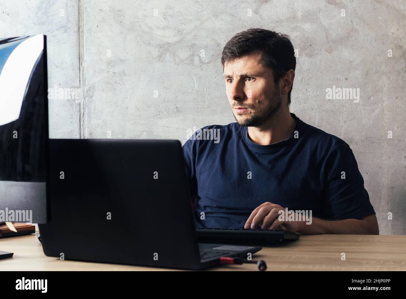Handsome serious bearded man sitting at the office desk, working on laptop, looking at computer screen against concrete wall in modern office Stock Photo