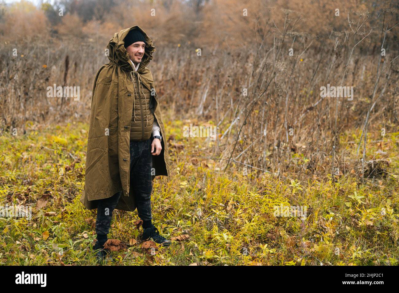 Portrait of smiling tourist man wearing green raincoat tent standing in thicket of bushes in cold overcast day and looking away. Stock Photo