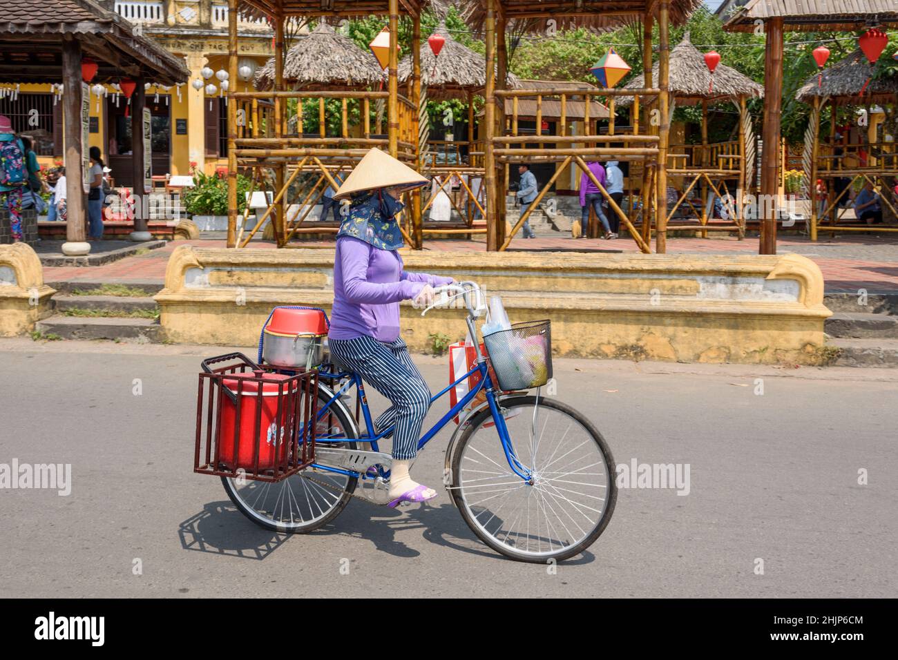 A Vietnamese woman wearing a traditional conical hat rides a bicycle through the streets of Hoi An, Quang Nam province, central Vietnam Stock Photo