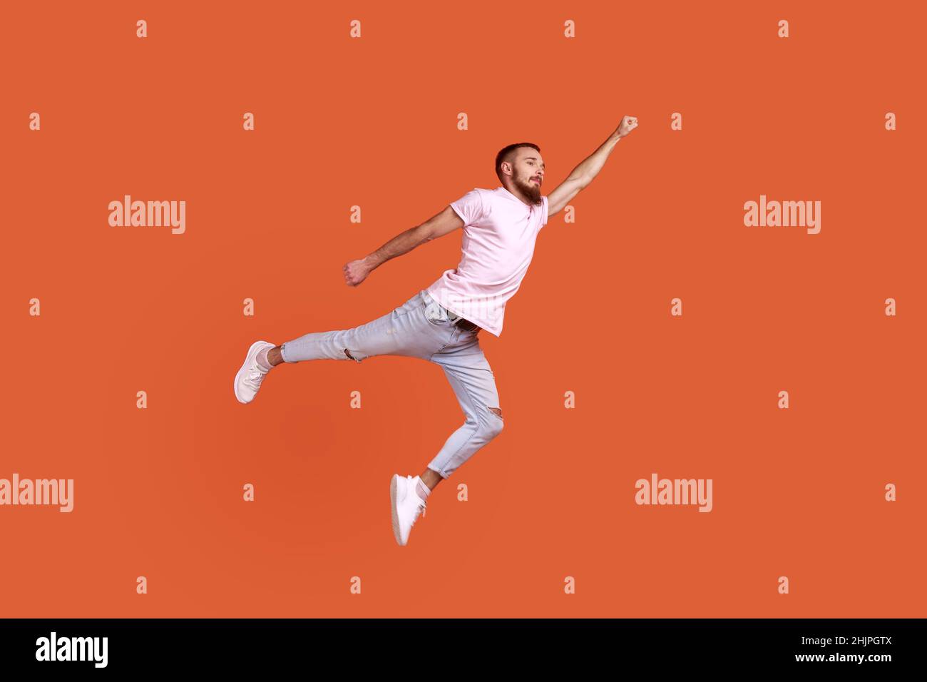 Portrait of motivated confident bearded man flying up in air as superhero with raised hand, feeling superpower, wearing pink T-shirt. Indoor studio shot isolated on orange background. Stock Photo