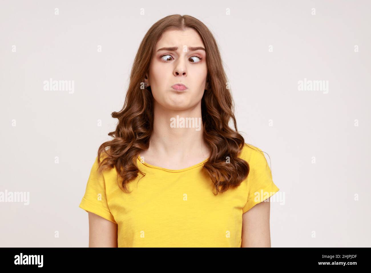 Portrait of attractive funny silly young woman in yellow T- shirt with cross eyed, has stupid dumb face, awkward confused comical expression. Indoor studio shot isolated on gray background. Stock Photo