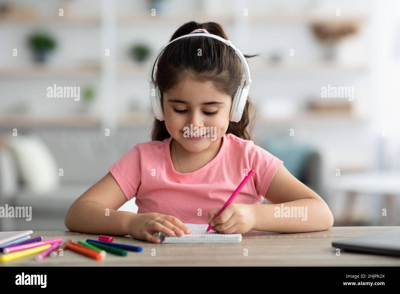 Kids Leisure. Little Arab Girl Wearing Wireless Headphones Drawing At Home Stock Photo