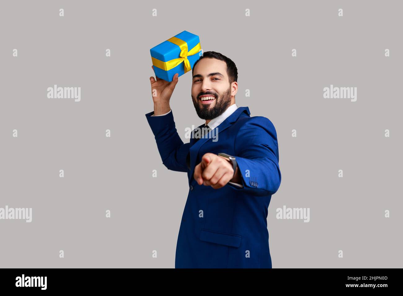 Portrait of smiling positive bearded man being ready to throw birthday box, having fun and sharing gift, wearing official style suit. Indoor studio shot isolated on gray background. Stock Photo