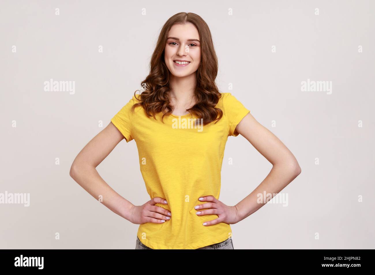Young female with brown wavy hair posing with hands on hips and looking directly at camera with positive and happy expression, wears yellow T-shirt. Indoor studio shot isolated on gray background. Stock Photo