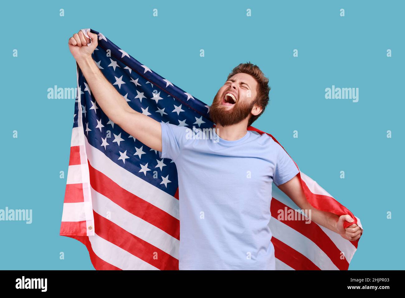 Portrait of delighted satisfied bearded man holing huge american flag and rejoicing while celebrating national holiday, looking up and yelling. Indoor studio shot isolated on blue background. Stock Photo
