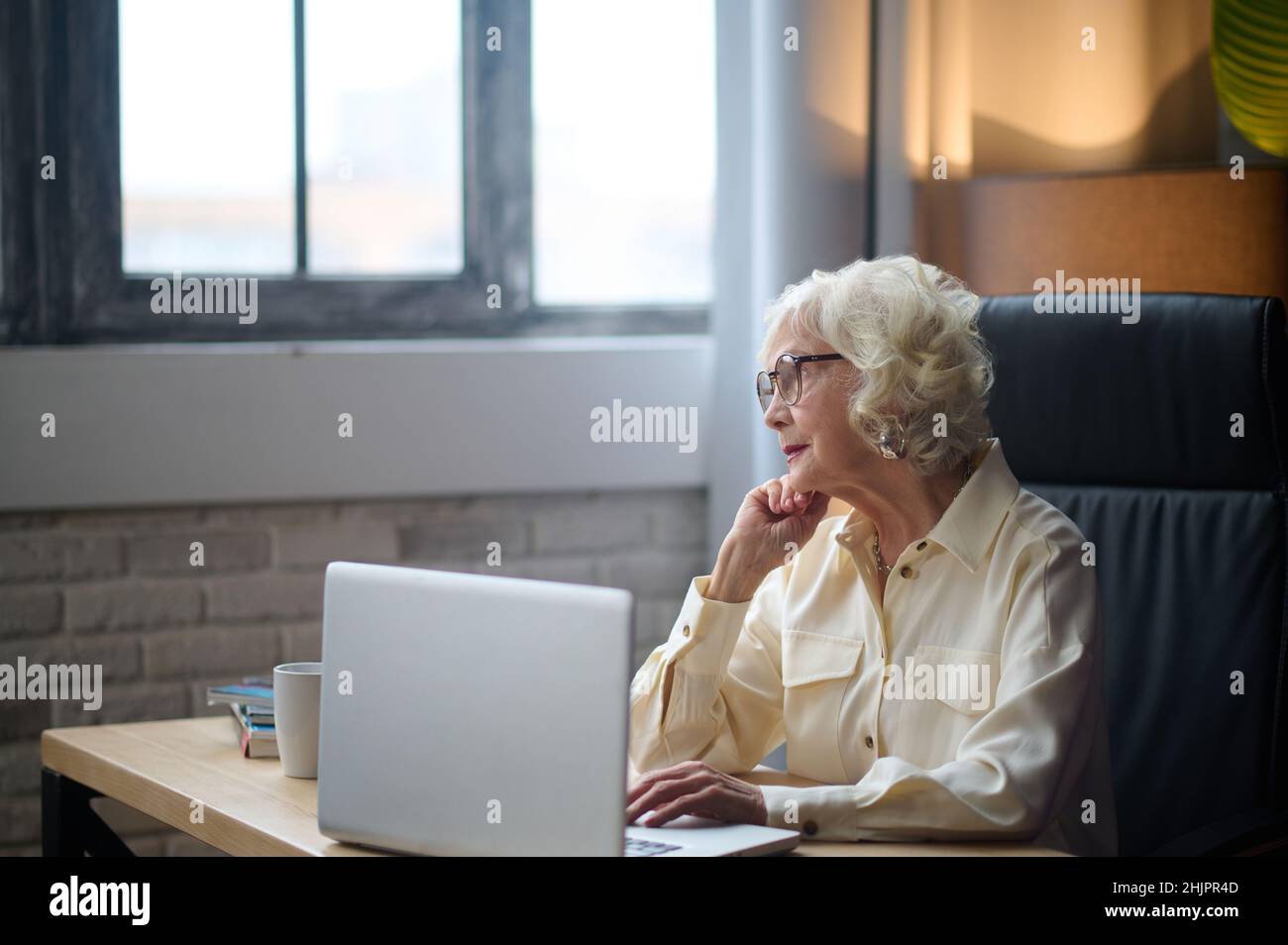 Woman looking at window sitting at table with laptop Stock Photo